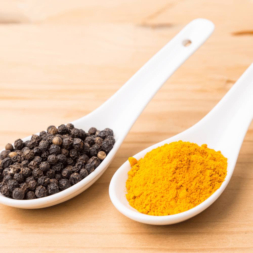 Two white spoons with black peppercorns and turmeric powder on a wooden surface.